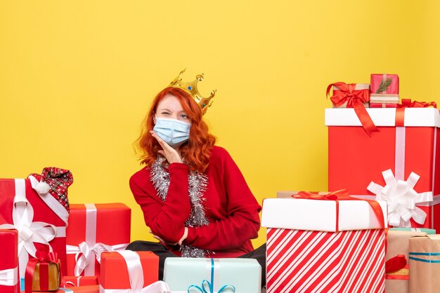 Front view of young woman sitting around presents in sterile mask on yellow wall