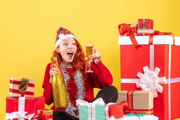 Front view of young woman sitting around xmas presents and celebrating with champagne on a yellow wall