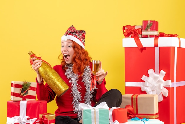 Front view of young woman sitting around xmas presents and celebrating with champagne on yellow wall