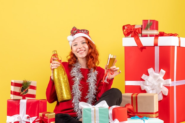 Front view of young woman sitting around xmas presents and celebrating with champagne on yellow wall