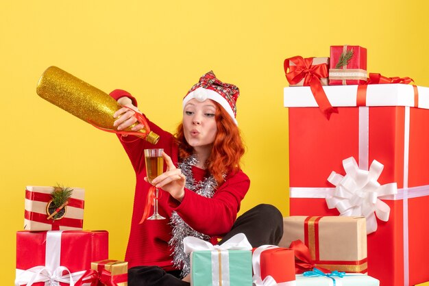 Front view of young woman sitting around xmas presents and celebrating with champagne on yellow wall