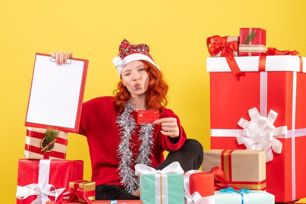 Front view of young woman sitting around xmas presents holding bank card on a yellow wall