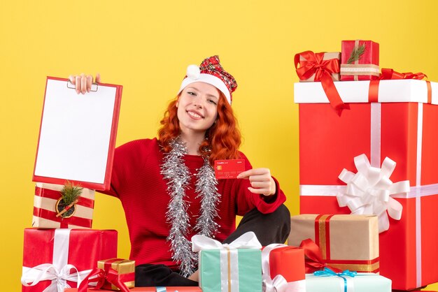 Вид спереди молодой женщины, сидящей вокруг рождественских подарков, держит банковскую карту на желтой стене