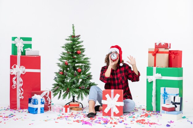 クリスマスプレゼントの周りに座っている若い女性の正面図は白い壁に時計を保持しています