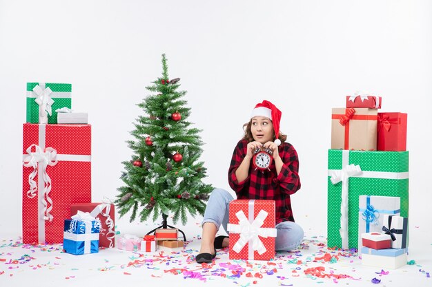 クリスマスプレゼントの周りに座っている若い女性の正面図は白い壁に時計を保持しています