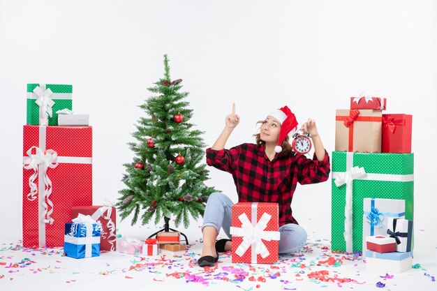 Front view of young woman sitting around xmas presents holding clocks on a white wall