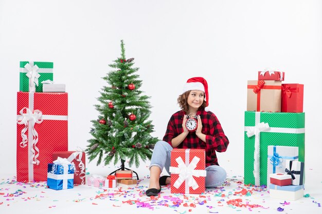 Front view of young woman sitting around xmas presents holding clocks on the white wall