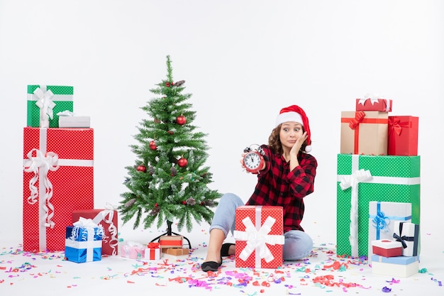 Front view of young woman sitting around xmas presents holding clocks on the white wall