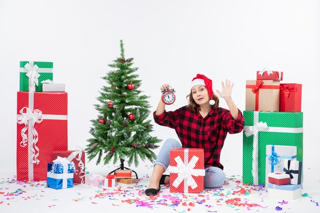 Front view of young woman sitting around xmas presents holding clocks on the white wall