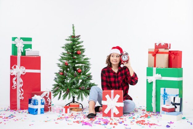 クリスマスプレゼントの周りに座っている若い女性の正面図は白い壁に時計を保持しています