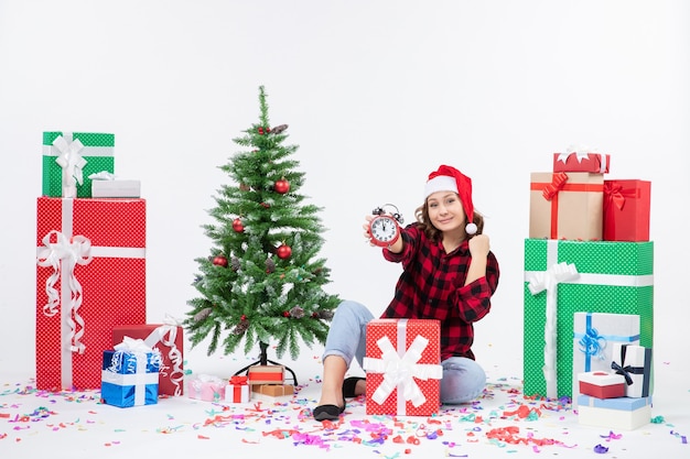 Front view of young woman sitting around xmas presents holding clocks on white wall