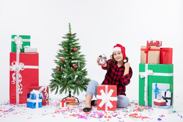 Вид спереди молодой женщины, сидящей вокруг рождественских подарков, держит часы на белой стене