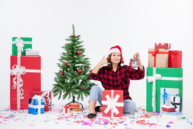 크리스마스 주위에 앉아있는 젊은 여자의 전면보기 흰 벽에 시계를 들고 선물