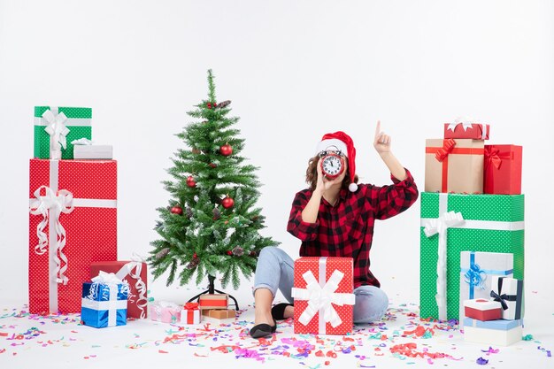 Front view of young woman sitting around xmas presents holding clocks on white wall