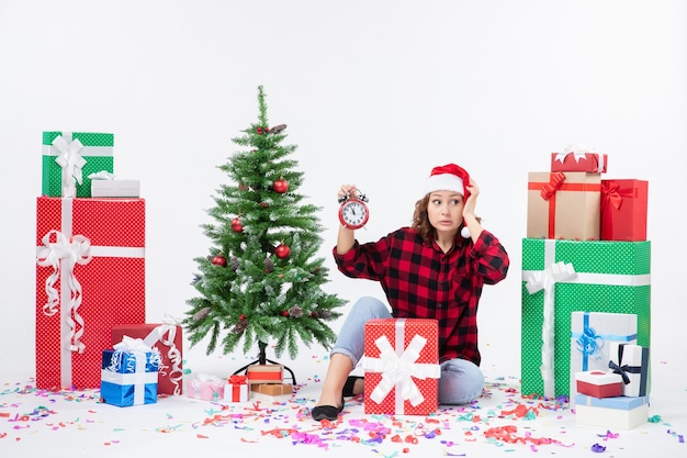 クリスマスプレゼントの周りに座っている若い女性の正面図は白い壁に時計を保持しています