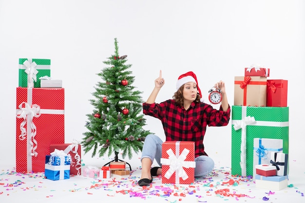 Front view of young woman sitting around xmas presents holding clocks on white wall