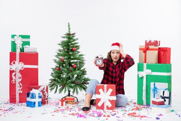 Front view of young woman sitting around xmas presents holding clocks on white wall