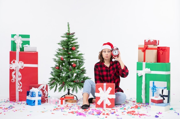 Вид спереди молодой женщины, сидящей вокруг рождественских подарков, держит часы на белой стене