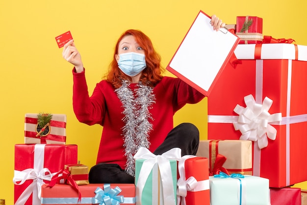 Free photo front view of young woman sitting around xmas presents in mask with bank card on a yellow wall