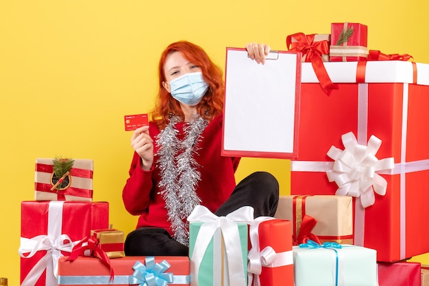 Front view of young woman sitting around xmas presents in mask with bank card on yellow wall