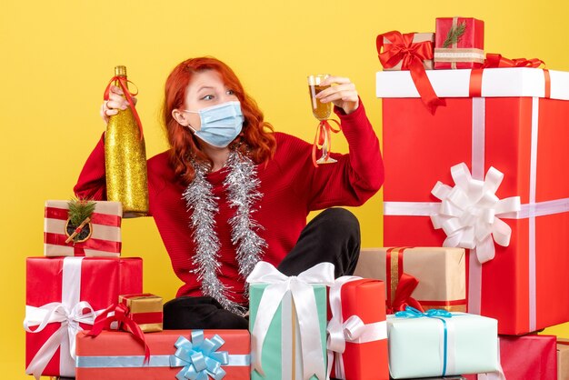 Front view of young woman sitting around xmas presents in mask with champagne on a yellow wall