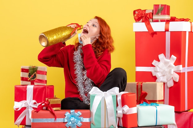 Vista frontale della giovane donna seduta intorno ai regali di natale in maschera con champagne sulla parete gialla