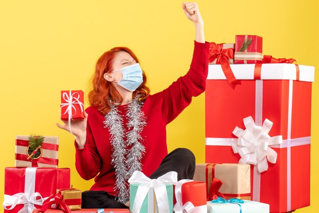 Front view of young woman sitting around xmas presents in mask on a yellow wall