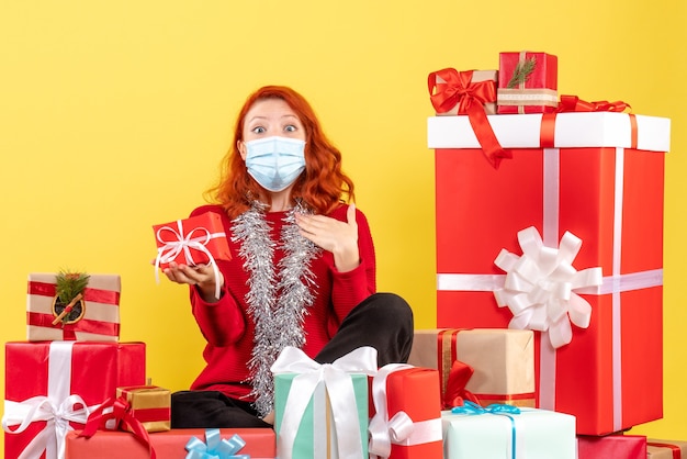 Vista frontale della giovane donna seduta intorno a regali di natale in maschera su una parete gialla