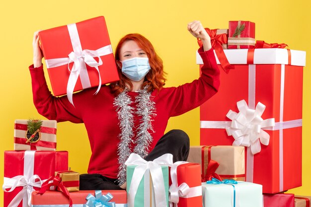 Front view of young woman sitting around xmas presents in mask on yellow wall