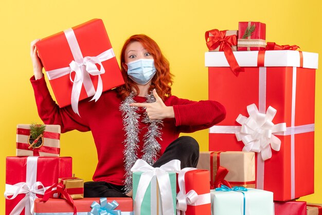 Front view of young woman sitting around xmas presents in mask on yellow wall
