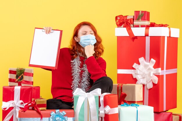 Вид спереди молодой женщины, сидящей вокруг рождественских подарков в маске на желтой стене