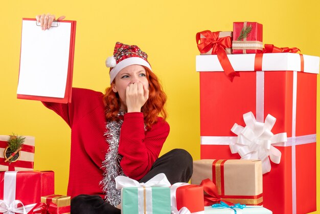 Front view of young woman sitting around xmas presents with file note on a yellow wall