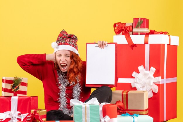 Front view of young woman sitting around xmas presents with file note on a yellow wall