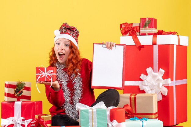 Front view of young woman sitting around xmas presents with file note on the yellow wall