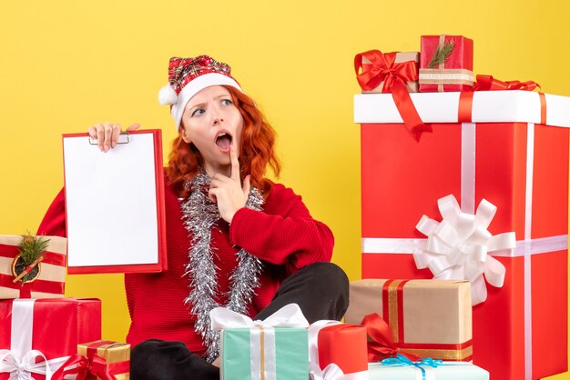 Вид спереди молодой женщины, сидящей вокруг рождественских подарков с файловой заметкой на желтой стене