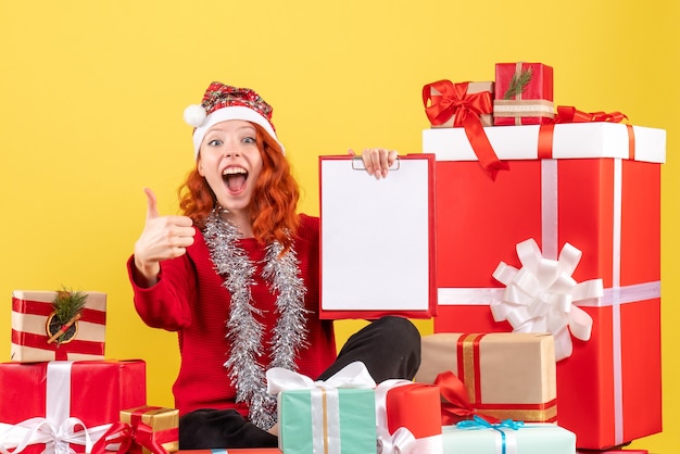 Front view of young woman sitting around xmas presents with file note on yellow wall