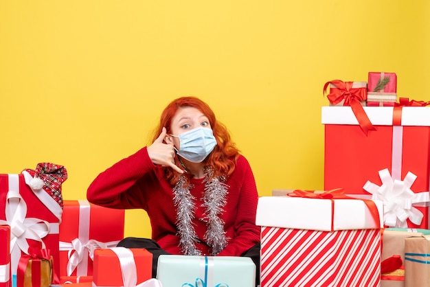 Вид спереди молодая женщина, сидящая вокруг рождественских подарков с маской на желтом новогоднем вирусе covid- color
