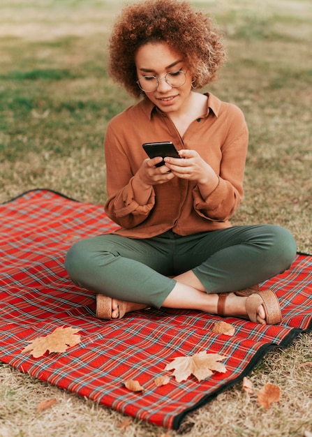 無料写真 彼女の電話をチェックしながらピクニック毛布の上に座っている正面の若い女性