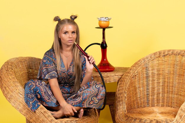 Front view of young woman sitting and smoking hookah on yellow wall