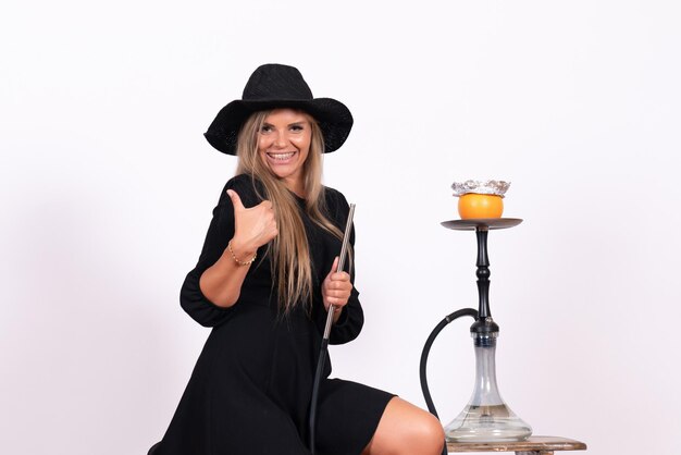 Front view of young woman smoking hookah and smiling on white wall