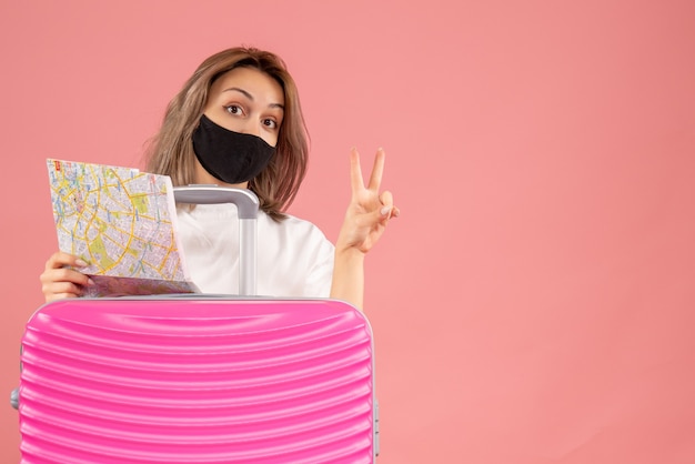 Vista frontale della giovane donna con la maschera nera che tiene la mappa che indica il segno di vittoria dietro la valigia rosa