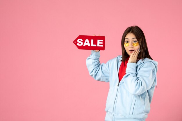 front view of young woman with eye patches holding red sale banner on pink wall
