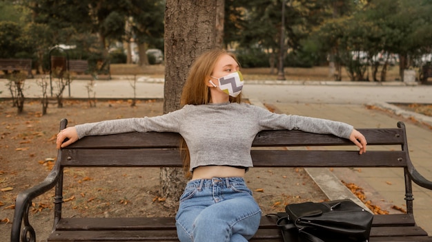 Foto gratuita giovane donna di vista frontale con mascherina medica che si siede su una panchina