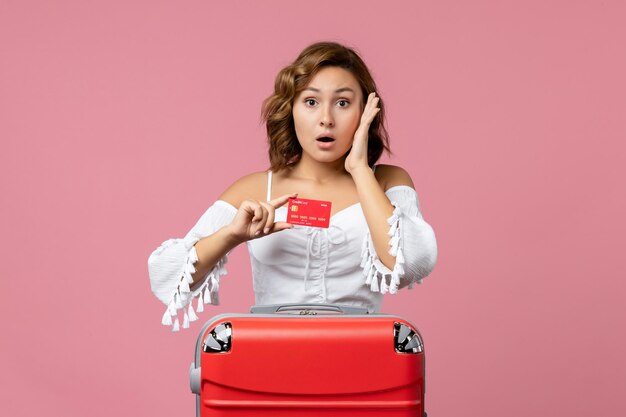 Front view of young woman with vacation bag holding bank card on pink floor vacation trip model voyage sea color