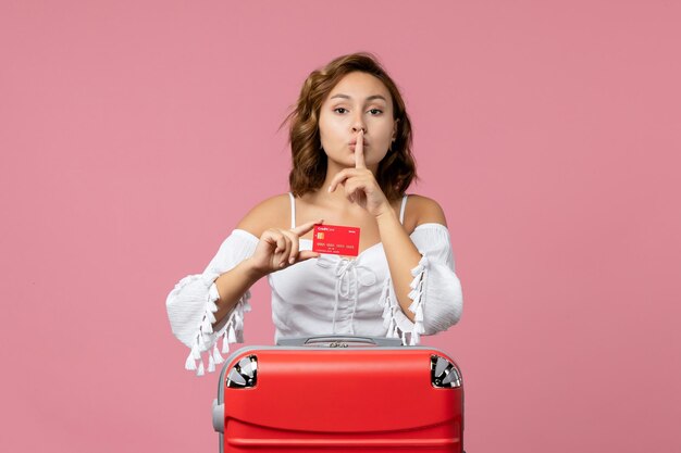 Front view of young woman with vacation bag holding bank card on pink wall