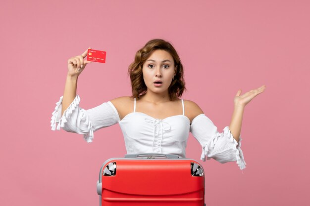 Front view of young woman with vacation bag holding bank card on pink wall