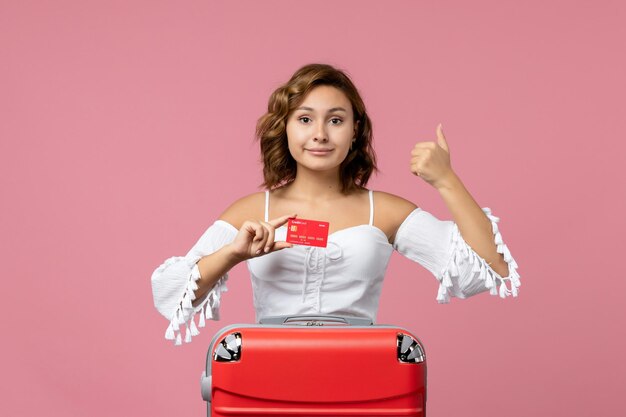 Вид спереди молодой женщины с туристической сумкой, держащей банковскую карту на розовой стене