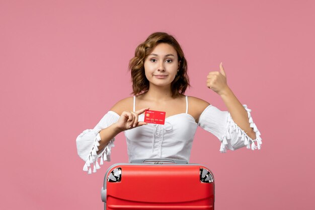 Front view of young woman with vacation bag holding bank card on the pink wall