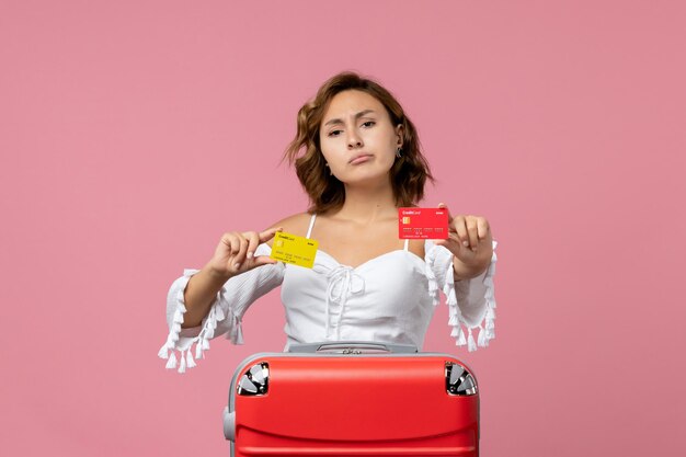 Front view of young woman with vacation bag holding bank cards on the pink wall