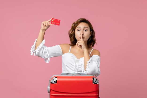 Вид спереди молодой женщины с туристической сумкой, держащей красную банковскую карту на розовой стене
